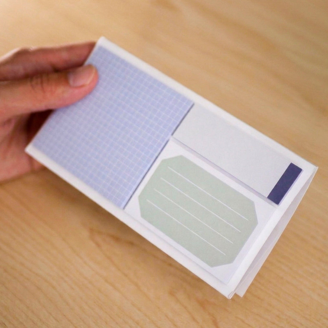 Hand holding a small sticky notepad with grid and plain sticky notes on a wooden surface