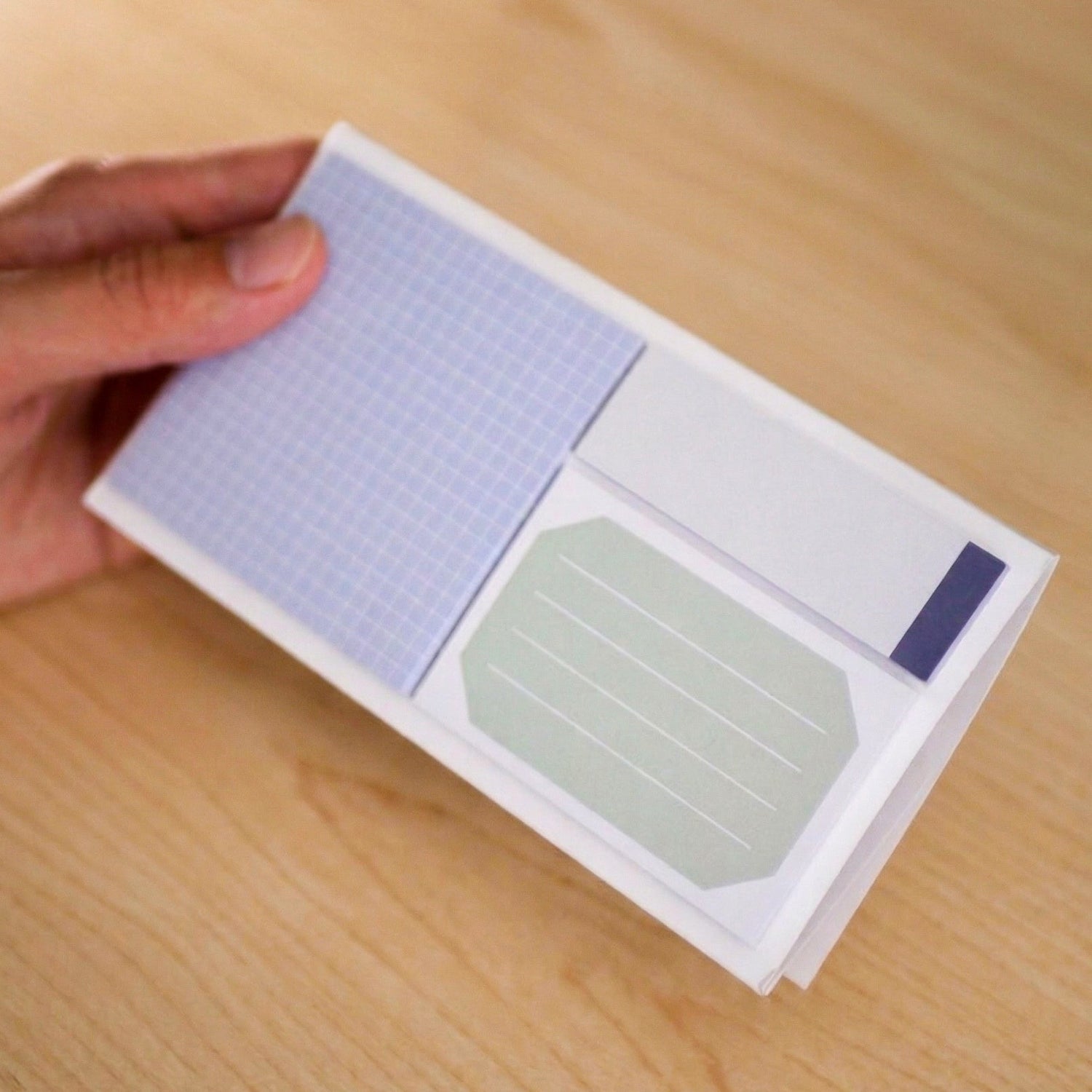 Hand holding a small sticky notepad with grid and plain sticky notes on a wooden surface